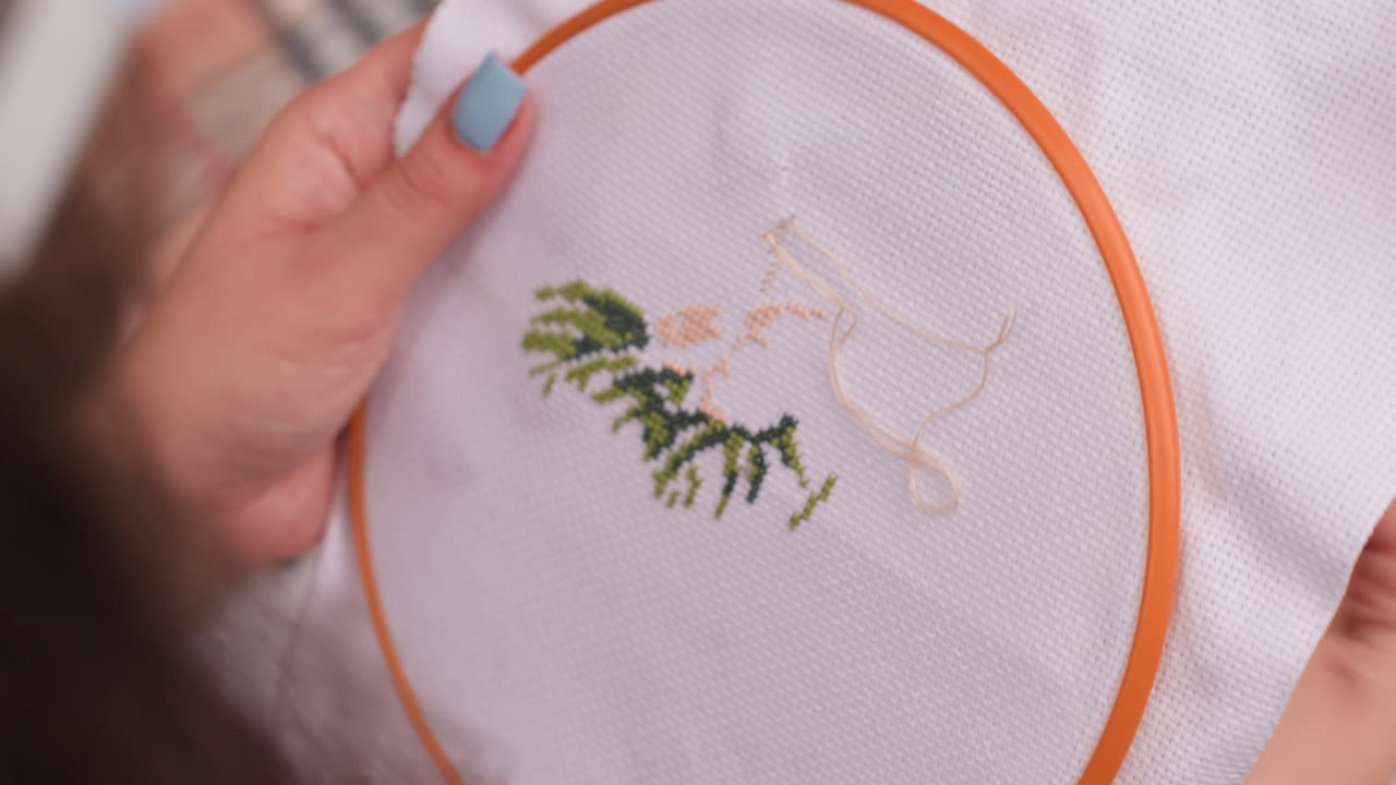 Close up of stylist with painted nails carefully embroidering fabric in orange hoop, finishing tree style cross stitch, patient hands guiding needle and thread under soft daylight
