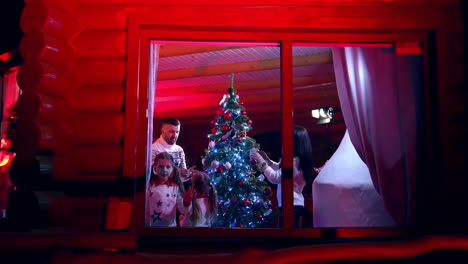 Family preparing for Christmas holidays.Parents and their children decorate New Year tree. Little girls looking through the window at Christmas eve. View from outside.