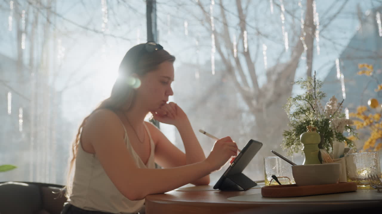 Lyst kreativt arbeid. Solskinn inspirerer til fokusert samarbeid i dag. Kvinne med nettbrett omgitt av livlig innredning. Fokusert kvinne i godt opplyst kafé engasjert i sin kreative planleggingsøkt