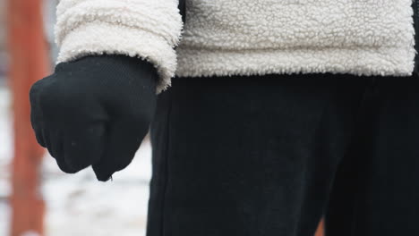 close-up of person wearing black gloves, winter jacket, and black trousers performing wrist twist, snow-covered ground and wooden workout equipment in background