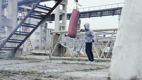 caucasian man in sportswear hitting a punching bag outdoors an abandoned factory on a cloudy morning