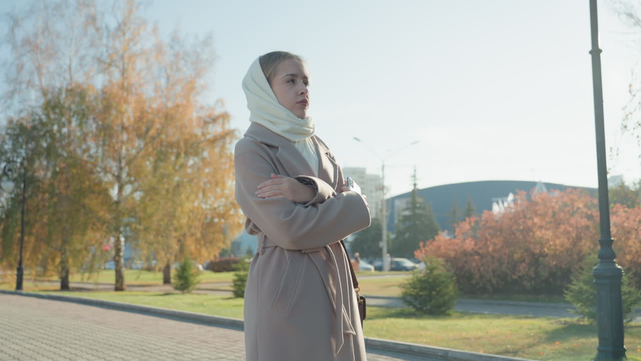 Kaukasische vrouw met hoofddoek in een herfstpark. Vrouw in jas klemt handtas en telefoon vast, wandelend onder gouden bomen. Stadsgezicht omlijst een student die haar telefoon checkt, een forens pauzeert bij een lantaarnpaal.