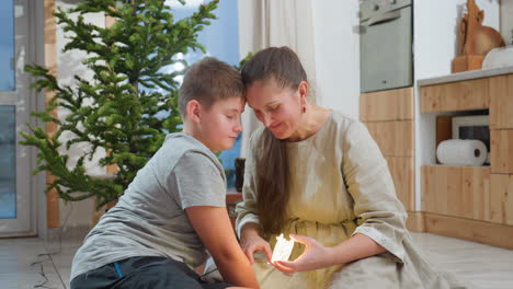 mom in grey gown and son in grey top and black shorts sit on floor in front of christmas tree smiling at each other while mom holds christmas light, sharing festive moments