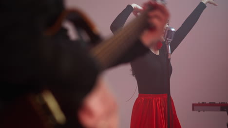 vocalist in a black and red gown performs passionately with a blurred guitarist's hand playing a red guitar in the background, creating a dynamic musical scene