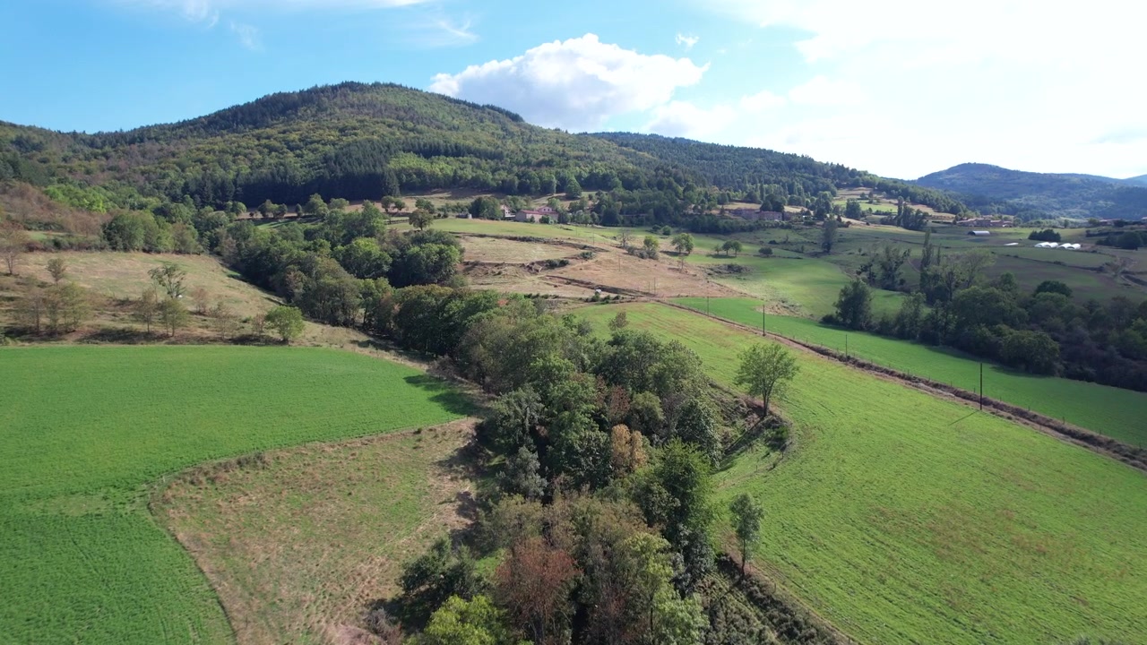 Uma tomada de drone em movimento mostrando a bela paisagem rural de Ardèche num dia claro. A vista apresenta campos exuberantes, árvores e colinas distantes, criando uma paisagem rural serena