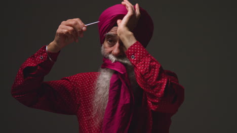 scatto di illuminazione in studio di bassa chiave di un uomo sikh anziano con la barba che lega tessuto per il turbante e utilizza l'ago salai contro uno sfondo scuro 1