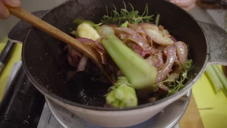 stirring beef shank mixed with onion slices, celery, spices with wooden spoon inside deep cooking pot