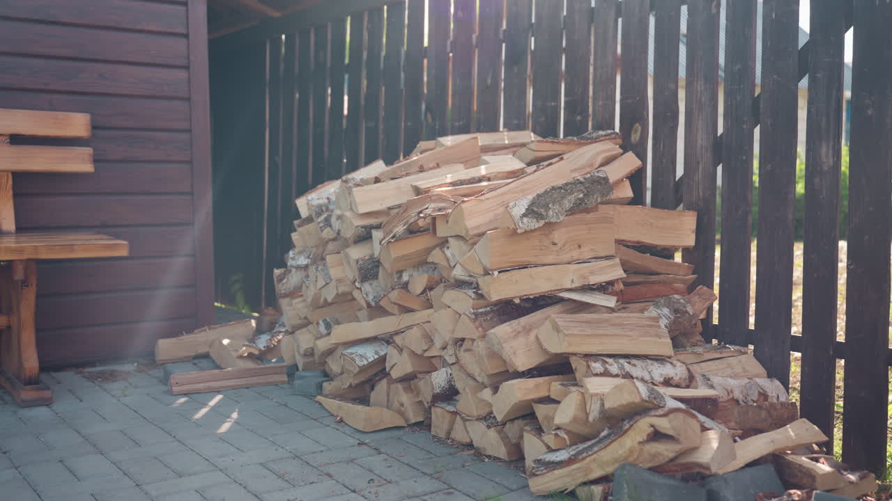 Stacked birch logs arranged neatly against dark wooden fence on paved ground with sunlight filtering through trees creating warm rustic ambiance for outdoor woodpile scene by cottage