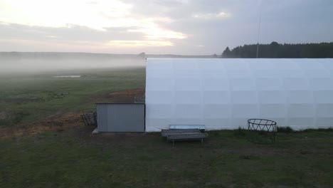 Weiße-Gewölbte-Halle-Im-Morgengrauen-Auf-Bauernhof-In-Polen-Sonnenaufgang-Luftaufnahme-Zirkulierende