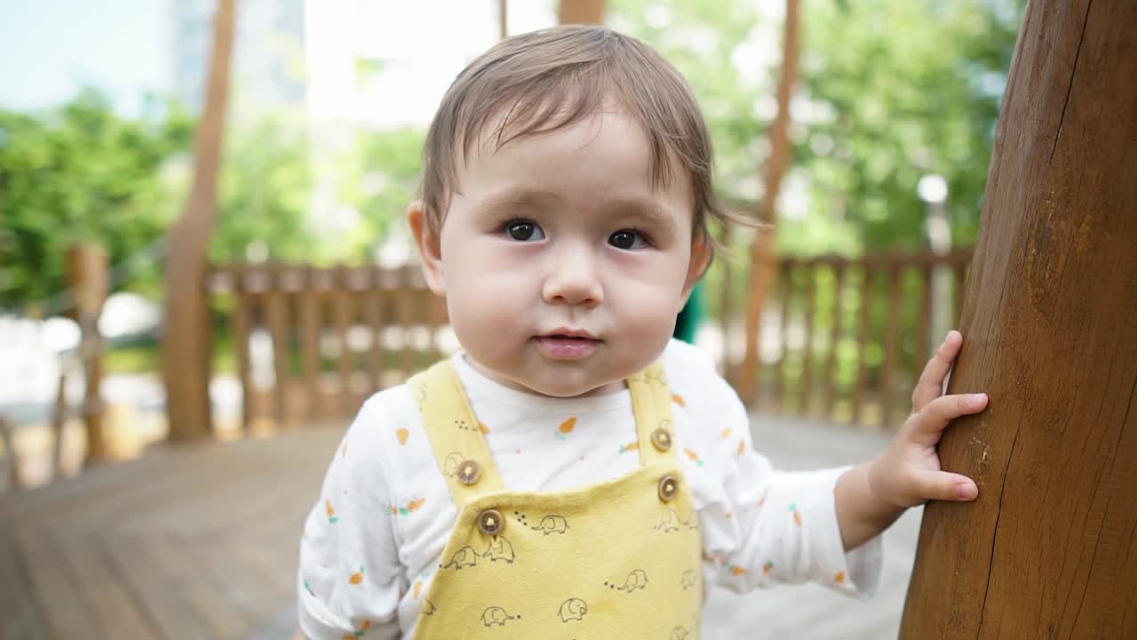 Attraktives, entzückendes Baby steht wackelnd von Seite zu Seite auf einem hölzernen Kinderspielplatz und blickt lieb mit einem leichten Lächeln in die Kamera.