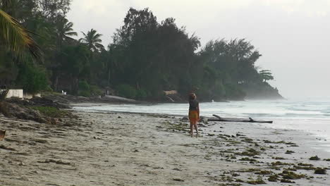 Una-Mujer-Camina-Por-Una-Playa-Agitando-Los-Brazos-Un-Perro-Corre-Y-Las-Olas-Se-Mueven