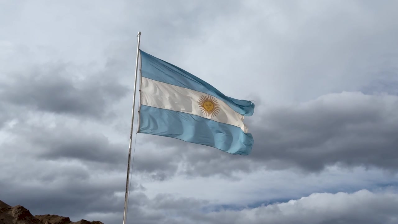 Drapeau argentin flottant au-dessus des contreforts andins sous des nuages d'orage spectaculaires et surplombant un paysage montagneux accidenté, plan fixe