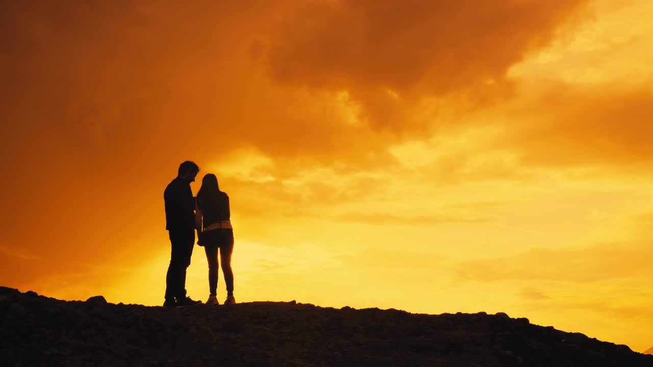 Silhouettes of young couple on mountain top enjoying orange yellow sunset