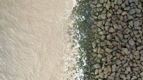 Sea-waves-hitting-rocks-on-a-sea-dyke,-drone-ascending-overhead-shot