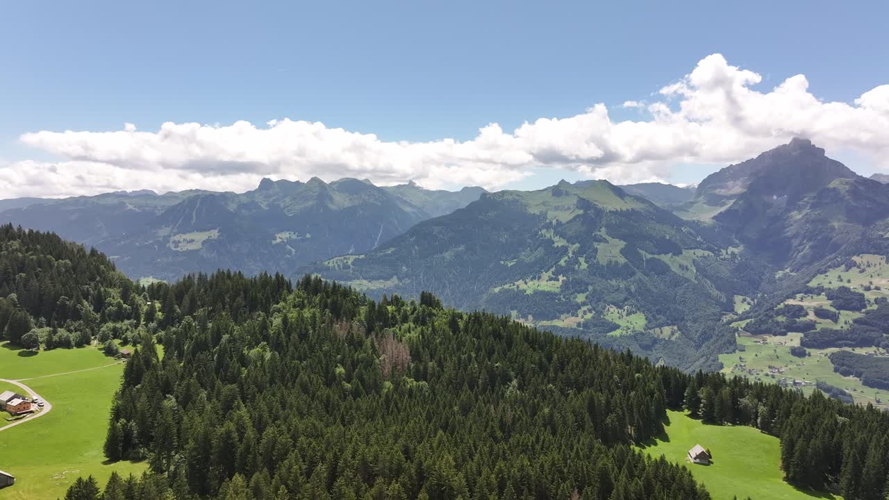 Drohnenflug mit Vorwärtsbewegung über üppig grünem Wald in den Schweizer Alpen, auf atemberaubende Berggipfel und blauen Himmel zufliegend.