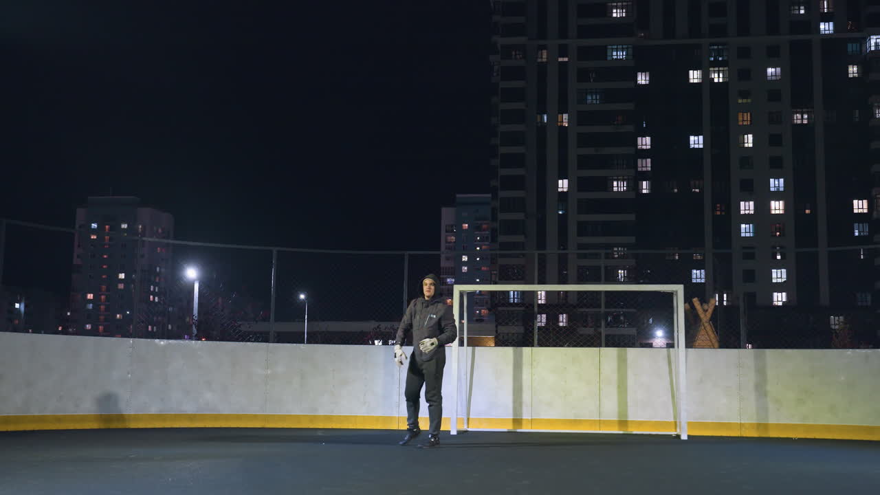 спортсмен готовится бросить футбольный мяч во время ночной тренировки возле ворота на освещенном городском поле, игрок демонстрирует спортивную форму под освещением стадиона с жилыми зданиями на заднем плане