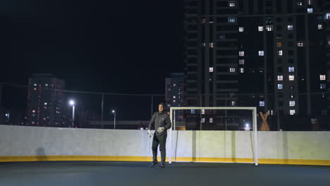 el atleta se prepara para lanzar una pelota de fútbol durante la práctica nocturna cerca del poste de la portería en un campo urbano iluminado, el jugador demuestra forma atlética bajo la iluminación del estadio con edificios residenciales en el fondo