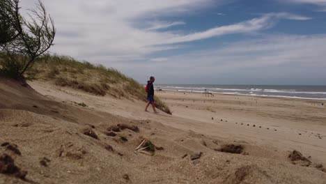 Acercar-La-Foto-De-Un-Hombre-Caminando-Entre-Dunas-De-Arena-A-Una-Playa-De-Arena-Durante-Un-Día-Ventoso---Mar-De-Las-Pampas,-Argentina