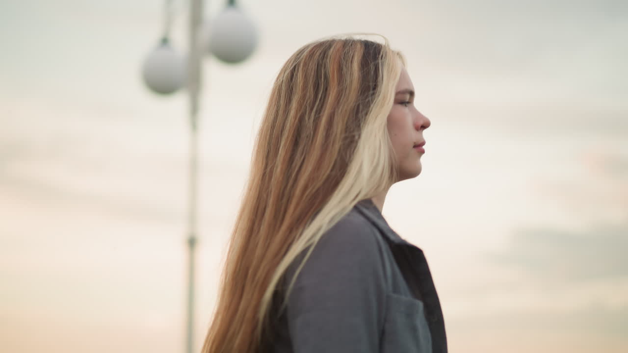 배경에서 두 개의 흐릿한 램프 기둥을 지나가는 여자의 근접 측면 뷰, 집중된 표정으로, 그녀는 긴 금발 머리카락을 가지고 있으며 부드러운 저녁 빛 아래 회색 옷을 입고 있습니다.