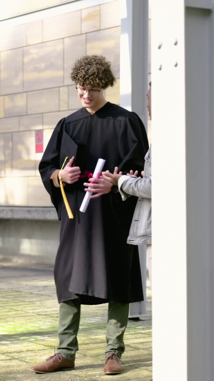Video verticale: Un laureato in toga che si allontana, tenendo in mano tocco e diploma, mentre un amico lo applaude su un viale del campus.