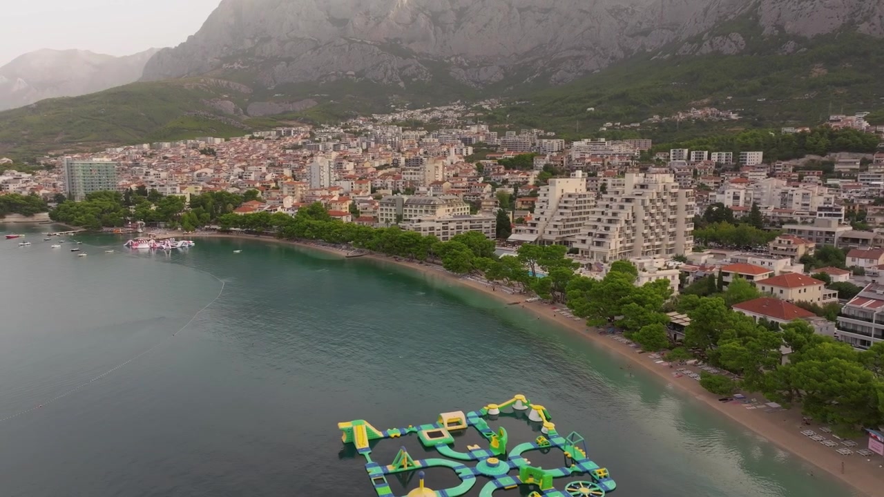 Parque aquático inflável flutuante posicionado próximo à praia arborizada de Makarska, com uma densa cidade litorânea e montanhas íngremes surgindo ao fundo