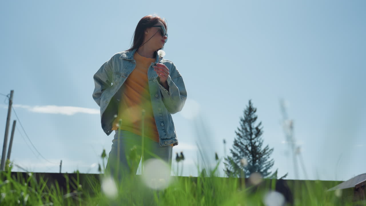 Young woman in denim jacket and sunglasses taking photo of dandelion using smartphone, standing in lush green grass with clear blue sky, tree, and fence in background