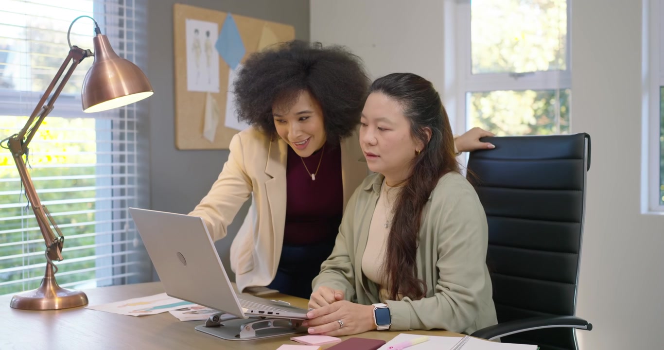 Diverse female team at desk using laptop, colleague entering, pointing and explaining, laughing