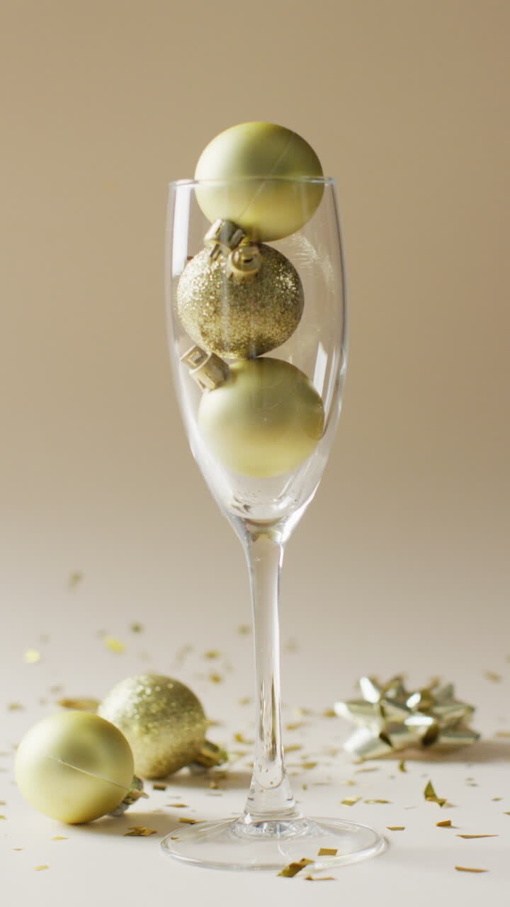 Vertical video of gold confetti falling over baubles in champagne flute glass