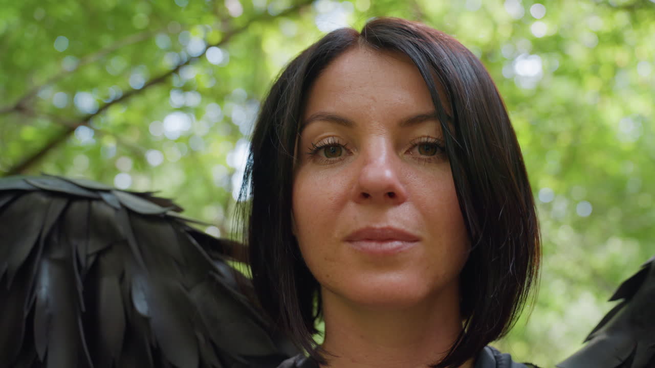 Close up of woman wearing black feather costume giving gentle air kiss with subtle smile in soft sunlight, surrounded by blurred green forest background creating dreamy atmosphere