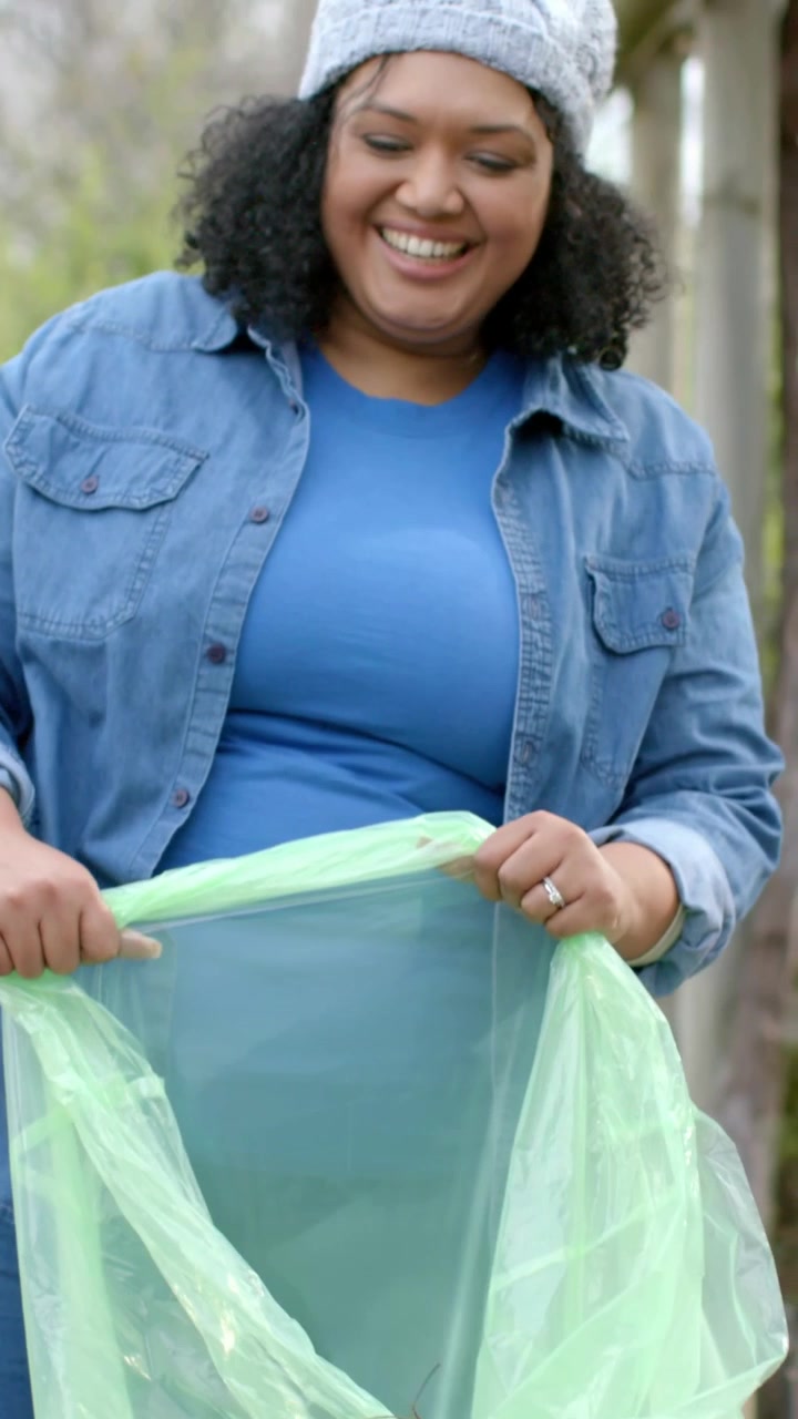 Vertical video: Holding woman in denim steadying bag to clean at park, gloved hand dropping twig