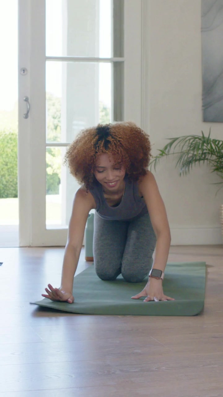 Vídeo vertical: Mulher com roupa de ginástica cinza começando yoga, desenrolando tapete verde em casa, com uma garrafa.