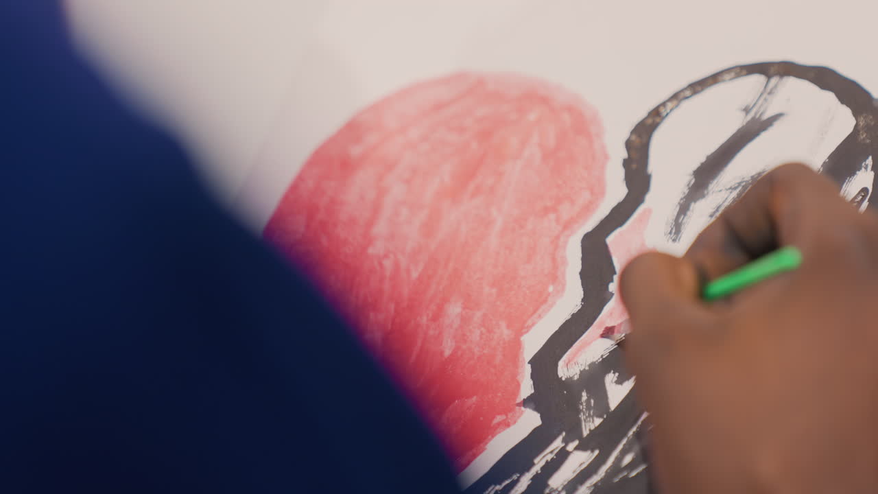 Tight frame hand outlining black painting, green brush tracing bold contour of red heart, shallow depth of field highlights textured pigment and deliberate line work, concentrated studio atmosphere