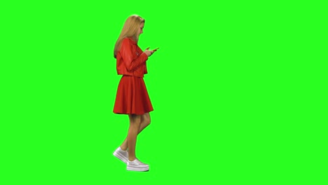 blonde girl is calmly walking and reading text message on her mobile phone at green screen. chroma key. profile view