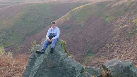 aerial footage circles a pensive young boy atop a substantial rock outcrop in the moorland, lost in thought