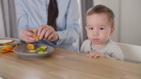 Unerkennbare-Mutter,-Die-Ihren-Kleinen-Jungen-Mit-Clementinensegmenten-Füttert,-Während-Sie-Zusammen-Am-Tisch-Im-Wohnzimmer-Sitzt