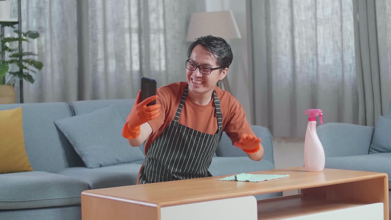 집에서 스프레이로 테이블을 청소하는 동안 스마트폰에서 비디오 통화를 하는 아프론을 입은 아시아 남성 집주인