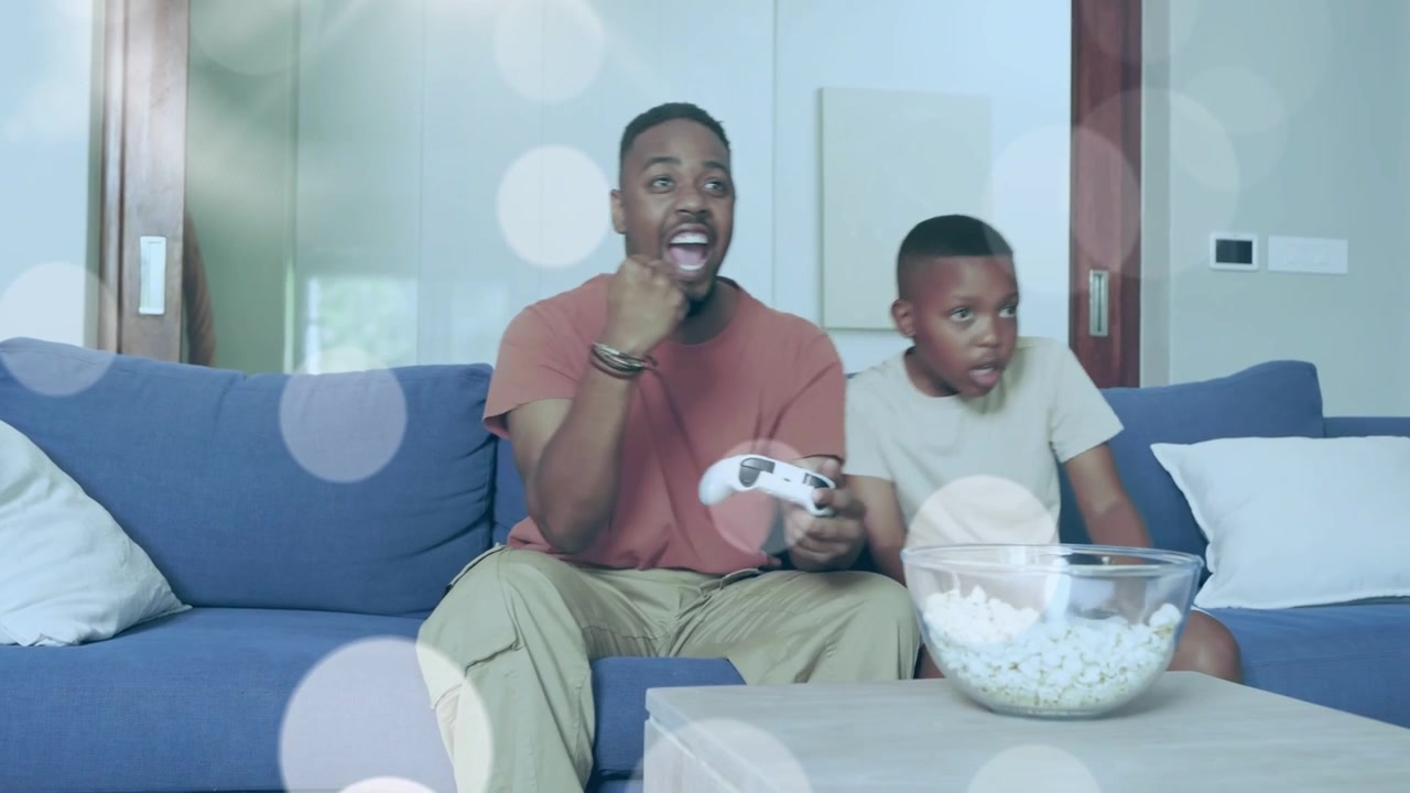 Far og søn med en controller reagerer på gaming-begivenheder og knytter bånd over en snack, midt i en scene sløret af bokeh.