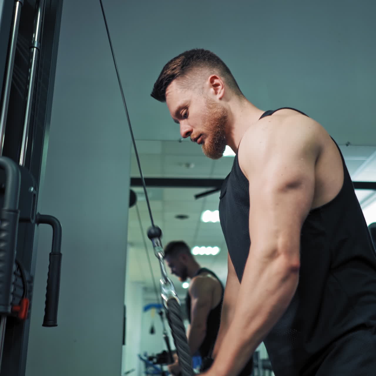 Muscular man trains on the simulator in fitness center. Strong athlete pull bar with rope and weights on fitness machine indoors.