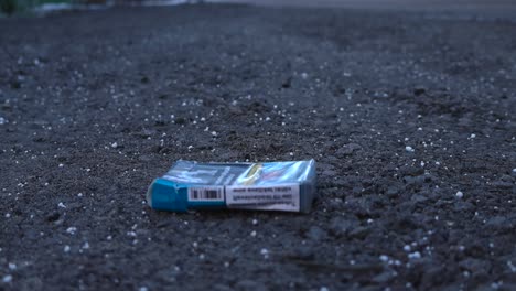 close up left to right panning footage of a blue and wrinkled cigarette pack that is on a dark gravel and muddy ground during a cloudy day. some snow grains are visible in the ground. in focus smokes