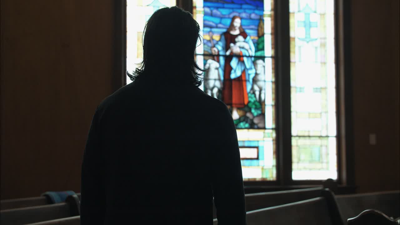 Back view of a man in dark clothing gazing at a stained-glass window, suggesting themes of redemption, purpose, and inner struggle
