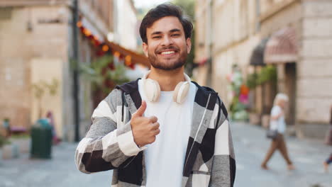 happy young indian man looking approvingly at camera showing thumbs up like gesture in city street