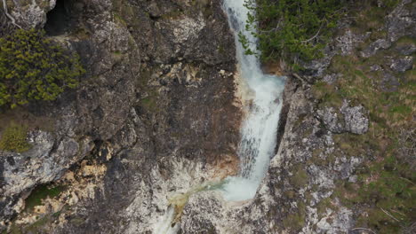 Aerial-close-up-view-of-a-waterfall-flowing-through-a-rocky-gorge-in-the-Dolomite-mountains,-surrounded-by-rugged-terrain