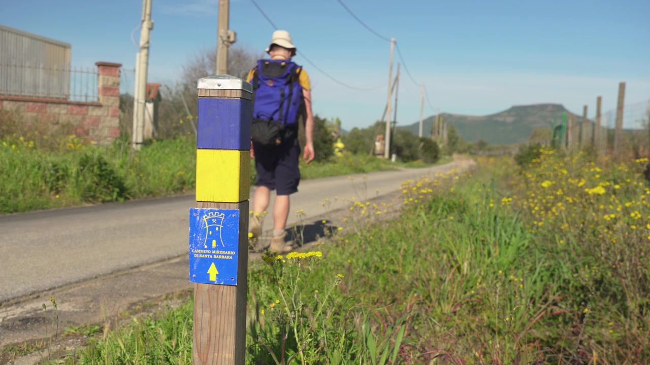 Man walking along the CMSB trail in Sardinia, Italy, showing the hiker on a natural path surrounded by rugged terrain and Mediterranean vegetation