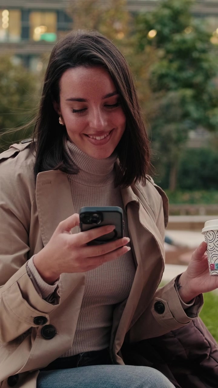 Giovane donna che usa lo smartphone e tiene in mano una tazza di caffè all'aperto