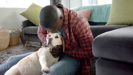 happy african american man sitting on floor at home, petting and kissing his pet dog, slow motion