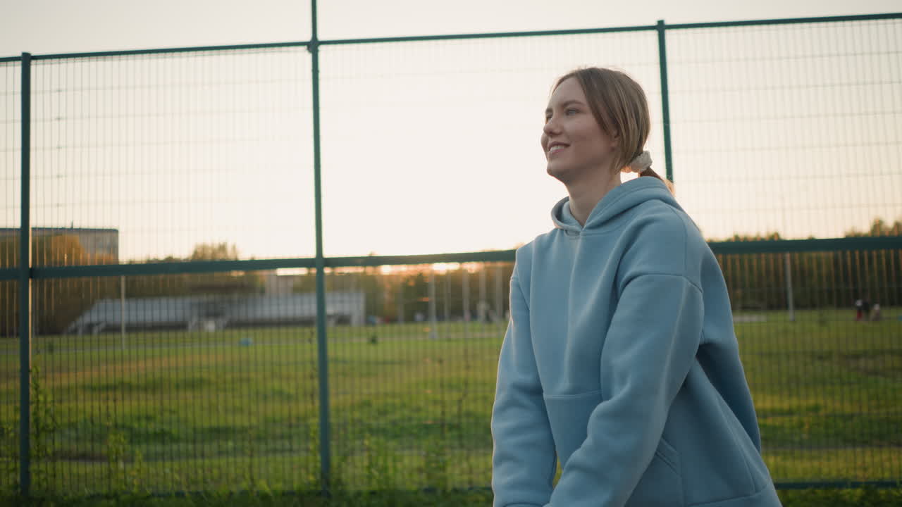 Junge Frau mit Kapuzenpullover lächelt während des Volleyballtrainings, spielt auf einem Outdoor-Feld mit einem warmen Lächeln und offenem Stadionhintergrund an einem sonnigen Tag