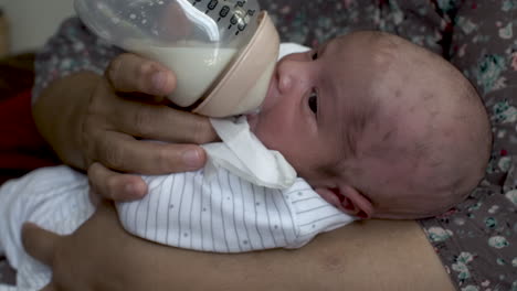 a close up shot of a newborn infant baby cradled in his mother’s arms feeding on a bottle of breast milk