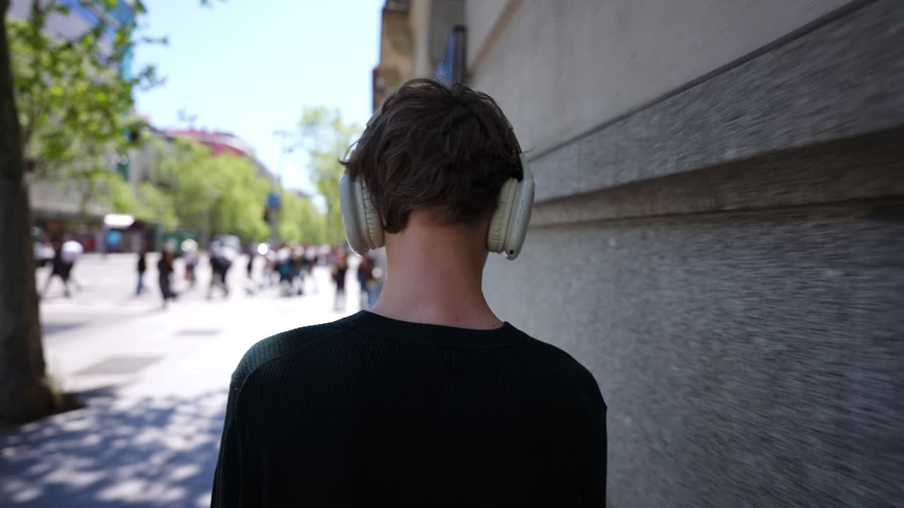 Teenager moving through european city center, enjoying music with wireless headphones, rear view capturing leisurely walking along scenic sidewalk during sunny day