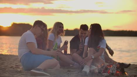 the group of young people is sitting in around bonfire on the sand beach. they are talking to each other and drinking beer at sunset.