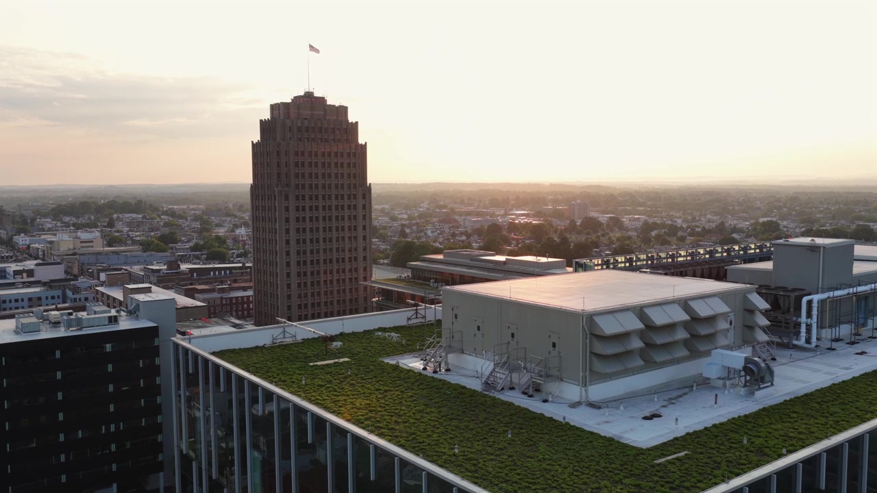 Jardim verde no telhado se estende ao lado de casa de máquinas e fileiras de venezianas de HVAC, torre Art Déco se eleva acima do horizonte nebuloso de Allentown ao pôr do sol dourado na Pensilvânia. Imagem aérea de drone.